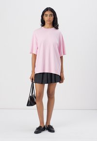 T-shirt oversized di cotone rosa chiaro con scollatura rotonda, indossata con una gonna a pieghe nera e scarpe nere, abbinata a una piccola borsa a mano nera.
