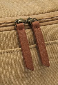 Tan canvas taske med bronze hardware og to brune faux læder lynlåse på toppen. Tekstureret stof med synlige sting.