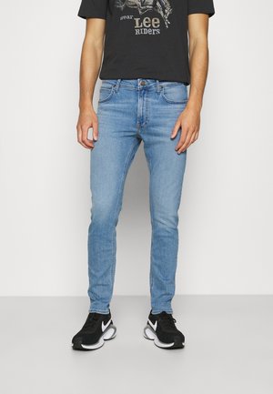 Man wearing light blue slim-fit jeans, black graphic tee with "Lee Riders" text, and black Nike sneakers standing against white background.