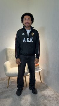 Uomo con capelli ricci che indossa una giacca nera A.C.K., jeans scuri e scarpe nere, in piedi sorridente in una stanza con una sedia bianca e un piccolo tavolo.