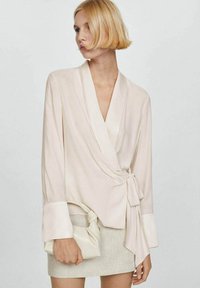 Blusa avvolgente beige chiaro con maniche lunghe, dettaglio in cravatta in vita e polsini a contrasto. Tessuto liscio con una leggera lucentezza.