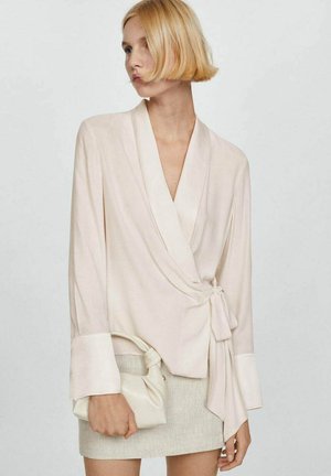 Blusa avvolgente beige chiaro con maniche lunghe, dettaglio in cravatta in vita e polsini a contrasto. Tessuto liscio con una leggera lucentezza.