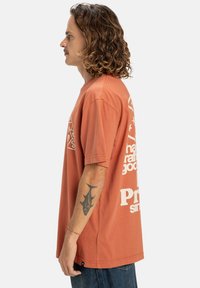 Hombre con cabello rizado, vistiendo una camiseta gráfica naranja y jeans azules, mostrando el perfil lateral y un tatuaje de pez en el antebrazo, de pie contra un fondo blanco.
