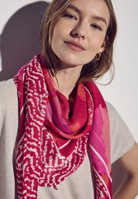 Helderrode en roze sjaal met abstracte patronen en een zachte textuur, om de nek gedrapeerd, met witte accenten en een zachte stof.