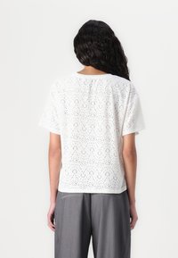 Top blanc à manches courtes avec des découpes au laser formant un motif. Coupe légèrement décontractée, tissu texturé, encolure arrondie, associé à un pantalon gris.