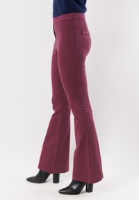 Pantaloni a zampa di elefante bordeaux realizzati in tessuto elastico, con vita alta, tasche frontali e una texture liscia, abbinati a stivali neri con tacco.