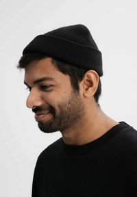 Zwarte gebreide muts met een aansluitend, taps toelopend ontwerp, gedragen door een persoon met een korte baard en een zwart shirt. Eenvoudige textuur, geen zichtbare patronen.