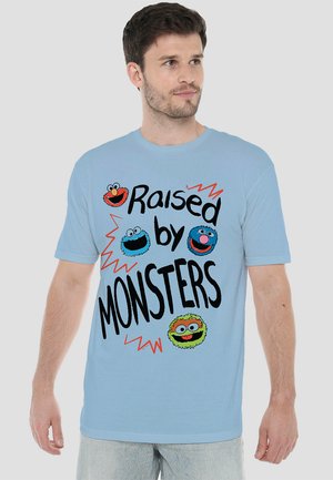 Helltürkises Baumwoll-T-Shirt mit kurzen Ärmeln, das bunte Charaktergrafiken und den Text "Raised by MONSTERS" in fetter schwarzer Schrift zeigt.