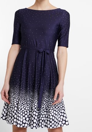 Marineblå kjole med korte ærmer, lille hvid prikket mønster, der overgår til større prikker nær den udvidede nederdel, bundet taljebælte.