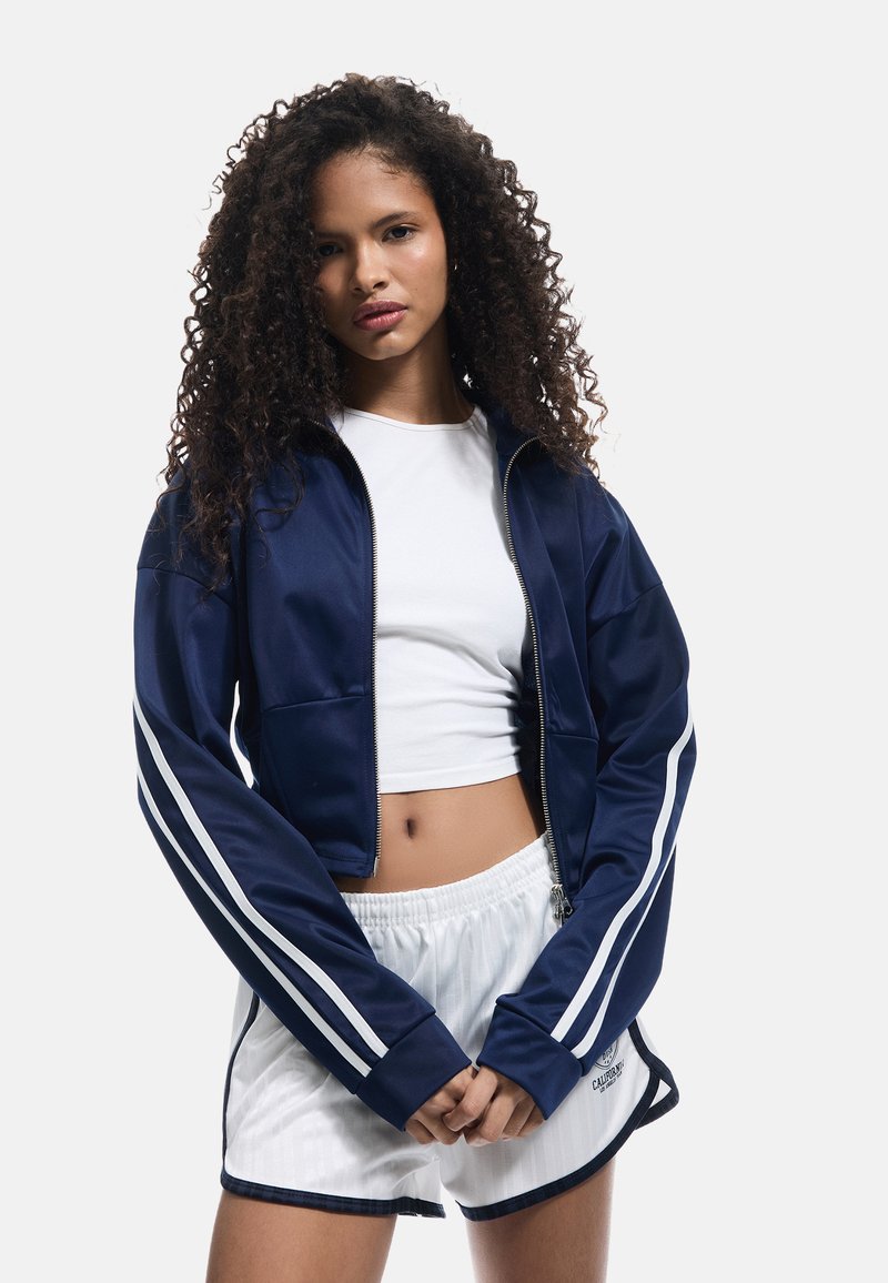 Mørkeblå zip-up jakke med hvide striber langs ærmerne, parret med en hvid cropped top og plisserede hvide shorts. Glat stof og atletisk pasform.