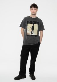 Gray cotton t-shirt featuring a graphic print of a silhouetted figure with the text "THE CURE BOYS DON'T CRY," paired with black pants and shoes.