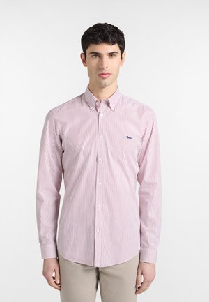 Ung mand iført en rød- og hvidstribet skjorte med lange ærmer og beige bukser, stående foran en ensartet hvid baggrund.