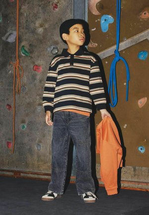 Gestreiftes Langarm-Polo mit blauen, beigen und orangenen Streifen; dunkle Jeans; schwarz-weiße Sneaker; hält einen orangefarbenen Kapuzenpullover. Kletterwand im Hintergrund.