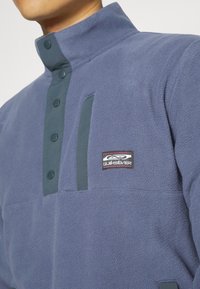 Fleece-pullover i mörkblå med hög krage, tryckknappsstängning och kontrasterande mörkgrå detaljer. Har en logotyp-patch på bröstet.