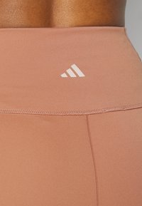 Gros plan d'une personne portant un legging marron clair taille haute avec un petit logo blanc Adidas sur la ceinture arrière.