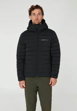 Sort puffer-jakke med hætte, med quiltet design, lynlåslukning foran og logo på venstre bryst. Kombineret med olivengrønne bukser.