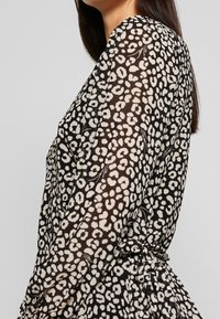 Blouse noire et crème avec un motif léopard sur un tissu transparent. Les manches longues présentent des détails froncés aux poignets.