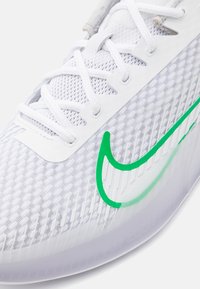 Bílé sportovní boty s texturovaným svrškem, zeleným logem Nike a viditelným systémem šněrování s průhlednou podrážkou.