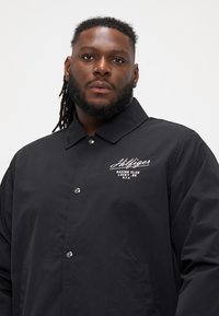 Crna windbreaker jakna s ovratnikom, kopčanjem na dugmadi i bijelim izvezanim logotipom na prsima. Glatka, mat završna obrada.