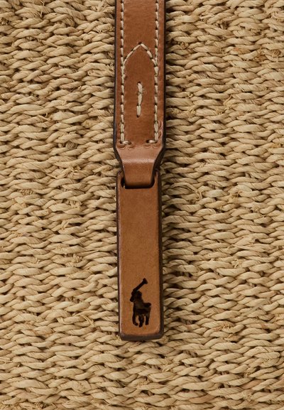 Närbild av ett flätat halmstruktur med en tanfärgad läderrem med vit söm och en liten svart polo spelare logotyp.
