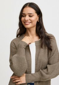 Cárdigan marrón de punto con mangas anchas y textura acanalada, llevado sobre una blusa blanca. El material parece suave y acogedor.
