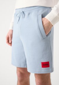 Shorts din bumbac de culoare albastru deschis, cu talie ajustabilă cu șnur, buzunare laterale și o eticheta rectangulară roșie cu logo-ul "HUGO" pe piciorul stâng.