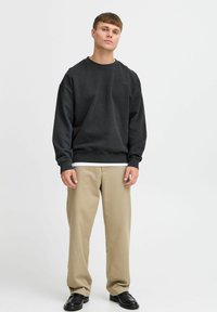 Must nounikaga dressipluus äärtega, kombineeritud beežide pükste ja mustade kingadega neutraalsel taustal.