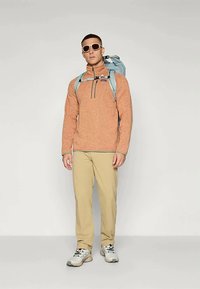 Moški v bež hlačah, oranžnem puloverju z zadrgo do četrtine, belih supergah, sončnih očalih in svetlo modrem pohodniškem nahrbtniku, ki stoji ob enobarvni steni.