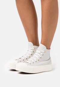 Hoge sneakers van lichtgrijs leer, met witte rubberen zolen, gouden veterogen en geaccenteerde textuur rond de enkel.