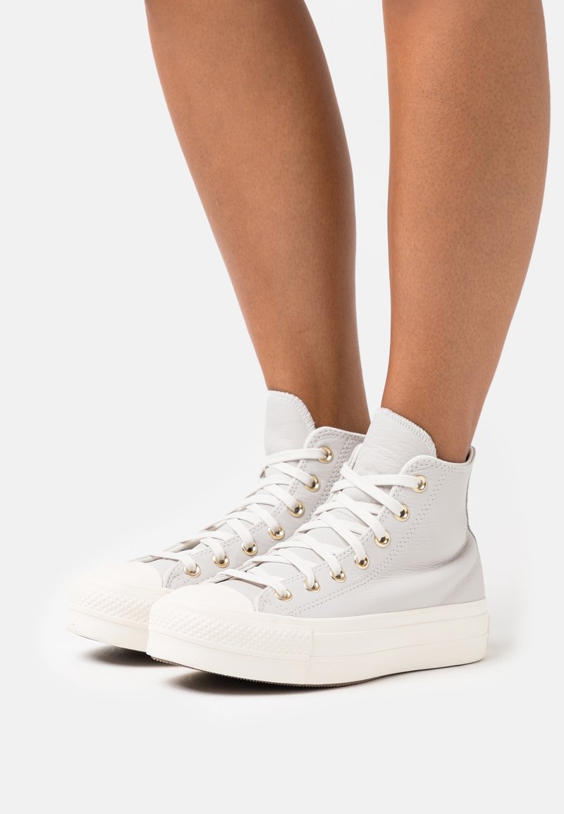 Hoge sneakers van lichtgrijs leer, met witte rubberen zolen, gouden veterogen en geaccenteerde textuur rond de enkel.
