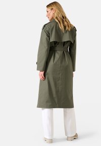 Manteau trench vert olive avec une taille ceinturée, un large col et un design à dos superposé. Associé à un pantalon blanc et des chaussures de couleur claire.