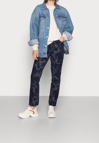 Chaqueta vaquera en azul claro, superpuesta sobre una sudadera color crema. Pantalones azul marino oscuro con un patrón tie-dye y zapatillas blancas gruesas.