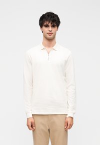 Langærmet, cremefarvet polo med lynlåskrave. Lavet af glat stof, den har et tætsiddende design og ribbede manchetter.