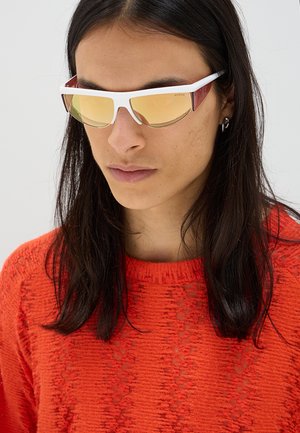 Persona con lunghi capelli scuri che indossa occhiali da sole riflettenti gialli con montatura bianca e un maglione strutturato arancione brillante su sfondo chiaro.