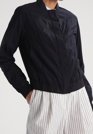 Persoon die een zwart jasje met rits draagt en een elastische taille, en witte broek met zwarte verticale strepen, staand met één hand in de zak.