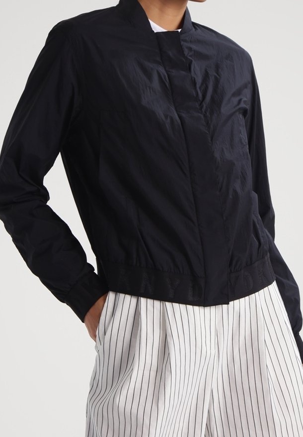 Personne portant une veste noire zippée avec une taille élastique et un pantalon blanc avec des rayures verticales noires, debout avec une main dans la poche.