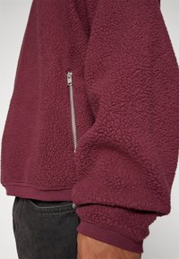 Sudadera de forro polar burdeos con una superficie texturizada y suave. Cuenta con un bolsillo con cremallera plateada en el lado izquierdo y puños y dobladillo acanalados.