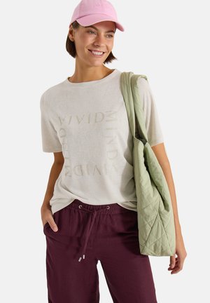 Smilende kvinde iført en beige T-shirt med grafik, vinrøde snøre bukser, lyserød baseballkasket og bærende en lysegrøn quiltet skuldertaske.