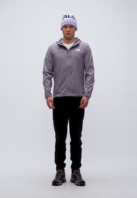 Άνδρας με γκρι μπουφάν με φερμουάρ, μαύρο παντελόνι, μποτάκια πεζοπορίας και λευκό σκουφάκι στέκεται μπροστά σε απλό λευκό φόντο.