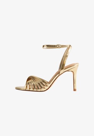 Gouden hoge hak sandaal met een gespschoen ontwerp, met een gedraaide voorkant, enkelband met gesp en een spitse neus. Gladde metallic afwerking.