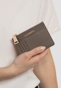 Cartera de cuero gris con superficie texturizada, múltiples ranuras para tarjetas, un tirador de cremallera dorado y el logo "LAZAROTTI" en relieve. Sostenida a mano contra un fondo blanco.