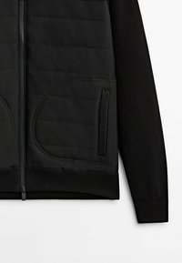 Veste noire à fermeture éclair avec panneau avant matelassé, poches latérales verticales lisses et manches longues en maille sur fond blanc.