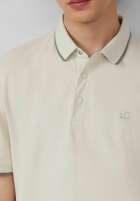 Beige Poloshirt mit Kragen, ausgestattet mit grauen Akzenten und einer Zwei-Knopf-Leiste. Ein kleines Logo ist auf der linken Brustseite sichtbar.