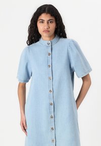 Lys blå denimkjole med korte puffermer, høy krage og full frontlukking med knapper. Har dekorativ søm og sidelommer.