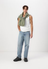 Hvit tanktop, grønn sweatshirt knyttet rundt skuldrene, lys blå dongeribukser og svarte joggesko. Modellen står foran en enkel bakgrunn.