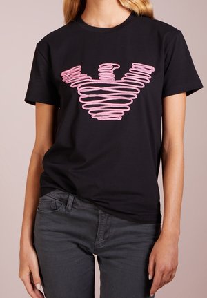 Zwart t-shirt met korte mouwen en een roze abstracte lijnontwerp op de borst dat lijkt op een gestileerde vogel of vleugels, gedragen met donkergrijze jeans.