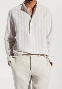 Hombre vistiendo una camisa de lino beige y blanca a rayas verticales con pantalones beige, con las manos en los bolsillos, frente a un fondo claro y liso.