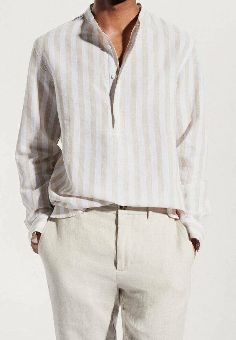 Hombre vistiendo una camisa de lino beige y blanca a rayas verticales con pantalones beige, con las manos en los bolsillos, frente a un fondo claro y liso.