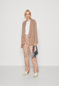 Béžový dlouhý blazer přes bílý top, vzorované volné kalhoty v růžové a krémové barvě, krémové podpatky, modrá taška se zlatými akcenty, stříbrný náhrdelník.