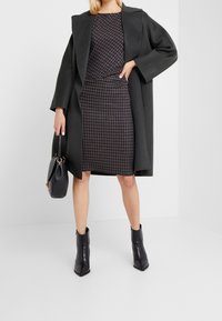 Donna che indossa un abito a quadri scuri, un lungo cappotto scuro, stivali neri alla caviglia e tiene una borsa nera, in piedi di fronte a uno sfondo bianco.